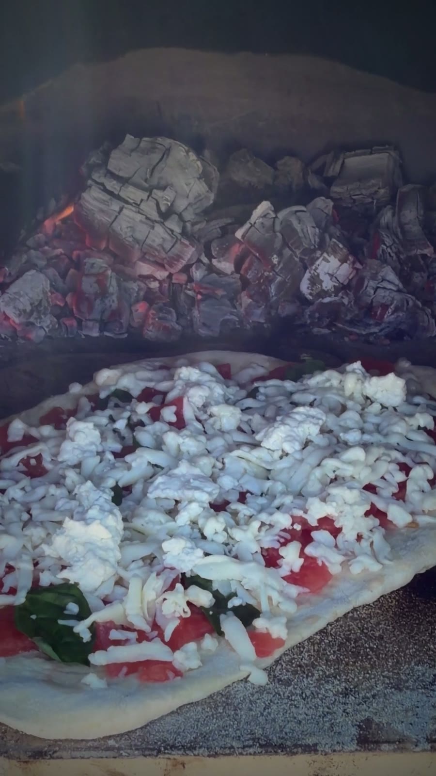 A pizza baking inside the wood-fired oven beside glowing coals